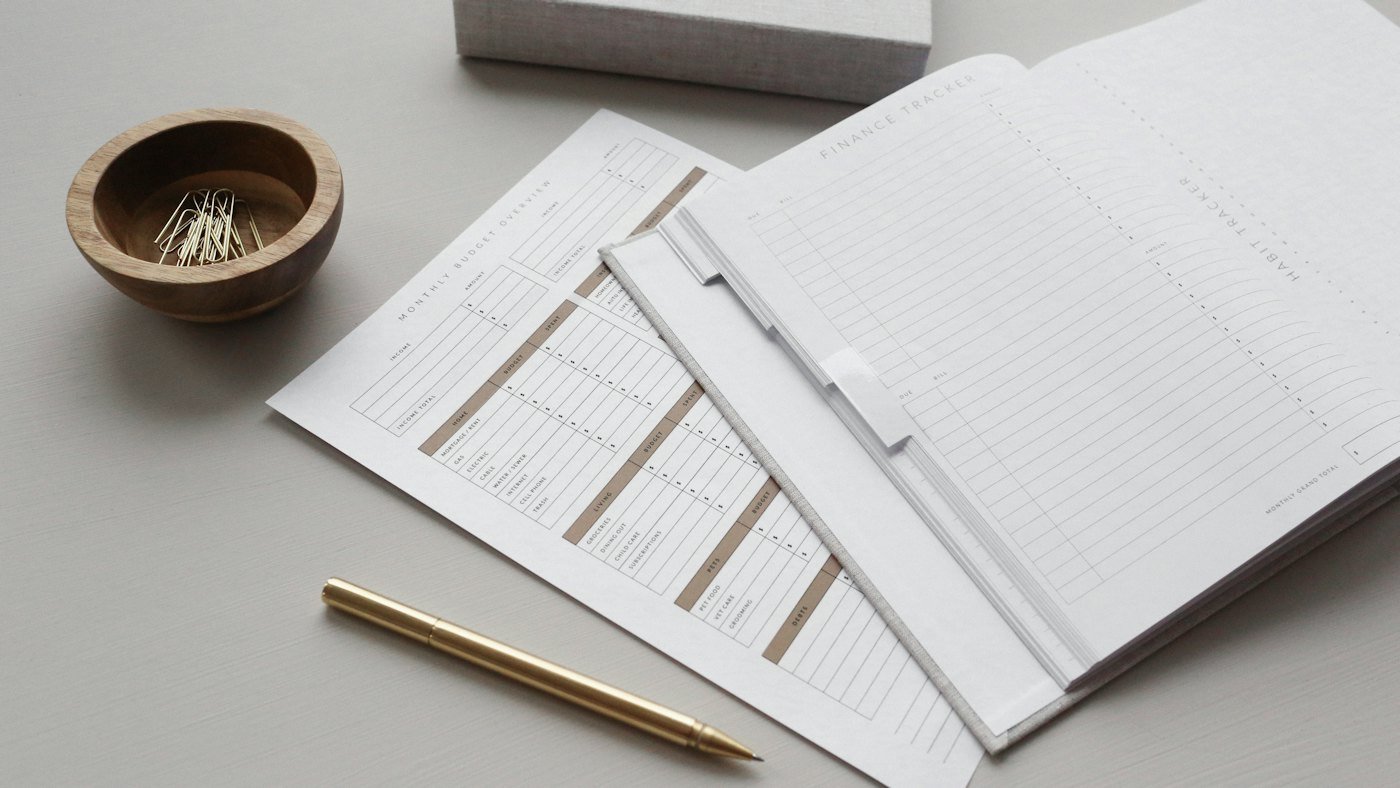 An open planner and a monthly budget sheet on a desk, with a pen and a bowl of paper clips