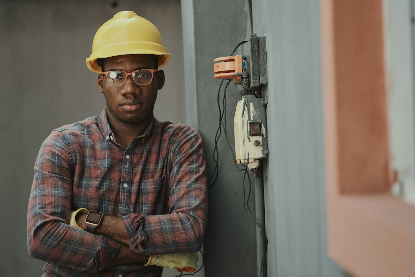 A self-employed electrician in a yellow hard hat standing with his arms folded