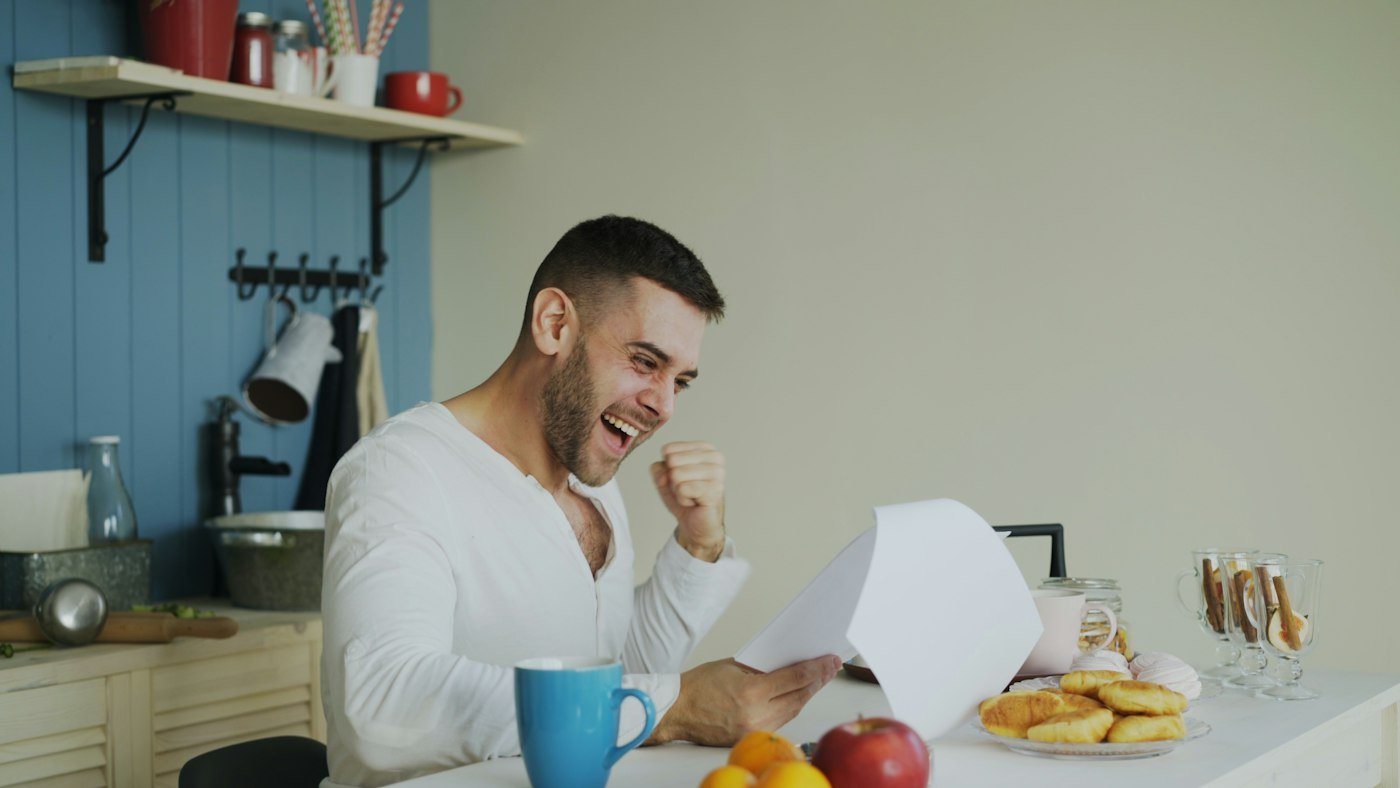 A man smiling as he reads a letter at his kitchen table